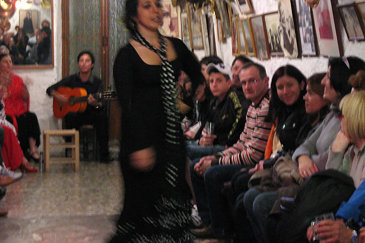 Mağaranın yan sıralarından izleyen seyircilerin önünde, puantiyeli atkılı siyahlar içindeki bir dansçı, oturan bir gitaristin yanından geçiyor