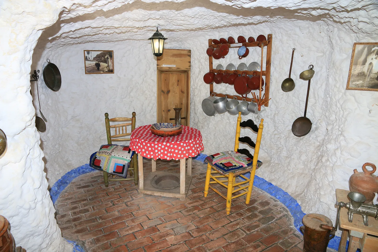 Ein weißgetünchter Höhlenraum mit einem rot gepunkteten Tisch, einem gelben Stuhl und Kupferpfannen an der Wand, nachgebaut im Sacromonte-Höhlenmuseum; ein allgemeines Foto eines Höhlenwohnsitzes, keine Schauhöhle